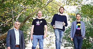 Four white men stand in front of trees, smiling. One is holding a gift box.