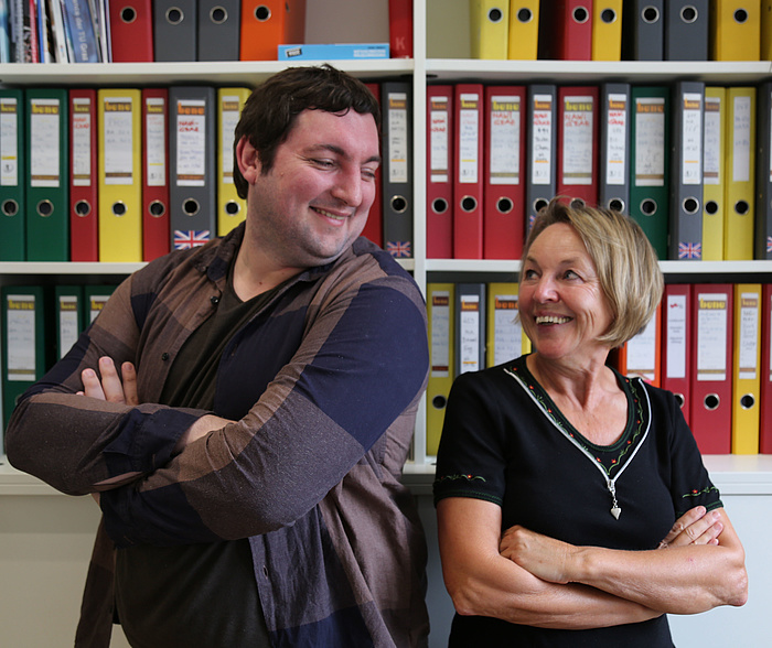 Christian Dobnik und Anna Maria Moisi lachen sich herzlich an - mit verschränken Armen Seite an Seite vor einer Wand von Aktenordnern im Büro an der TU Graz.