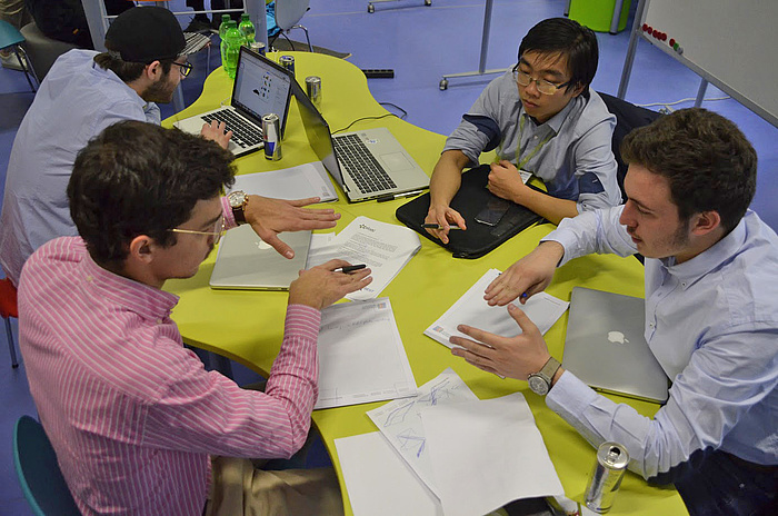 Vier Studenten sitzen mit Laptops und schriftlichen Unterlagen an einem knallgelben Tisch und diskutieren über die theoretische Problemlösung.)