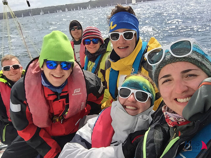 Equipped with sailing clothes, sunglasses, woolly hats and headbands, seven members of the HPS team smiling at the selfie camera on their boat.