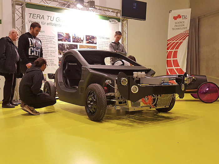 Ein IBEX-Modell vor einer Präsentationstafel des TERA TU Graz-Teams, umringt von Interessierten, die sich das Fahrzeug genauer ansehen.