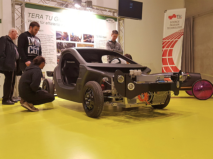 An IBEX model in front of a presentation board of the TERA TU Graz team, surrounded by interested persons looking at the vehicle in detail.