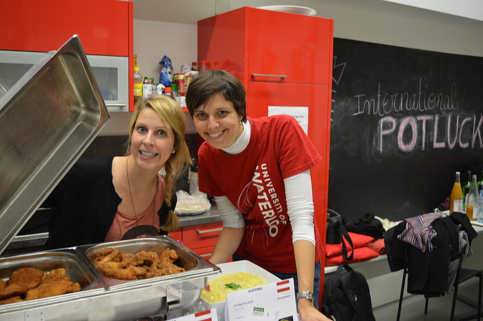 Zwei Mitarbeiterinnen des TU Graz Welcome Centers freuen sich am Duft von Wiener Schnitzeln in der Warmhaltepfanne und dem daneben angerichteten Kartoffelsalat.