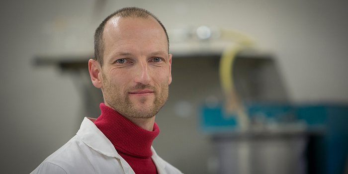 TU Graz-Wissenschafter Joachim Juhart vom Institut für Materialprüfung und Baustofftechnologie mit angeschlossener TVFA für Festigkeits- und Materialprüfung. Porträtfoto mit Labormantel und rotem Rollkragen.