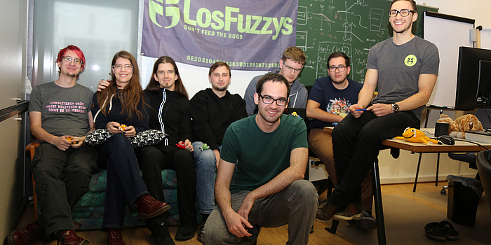 Acht LosFuzzys-Teammitglieder im Fuzzylab, im Hintergrund eine voll beschriebene Tafel und die violette LosFuzzys-Fahne.