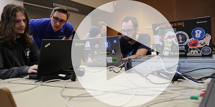 Three young men looking at two Laptops. In the middle of the picture you can see an arrow, meaning a Play-Button for videos.