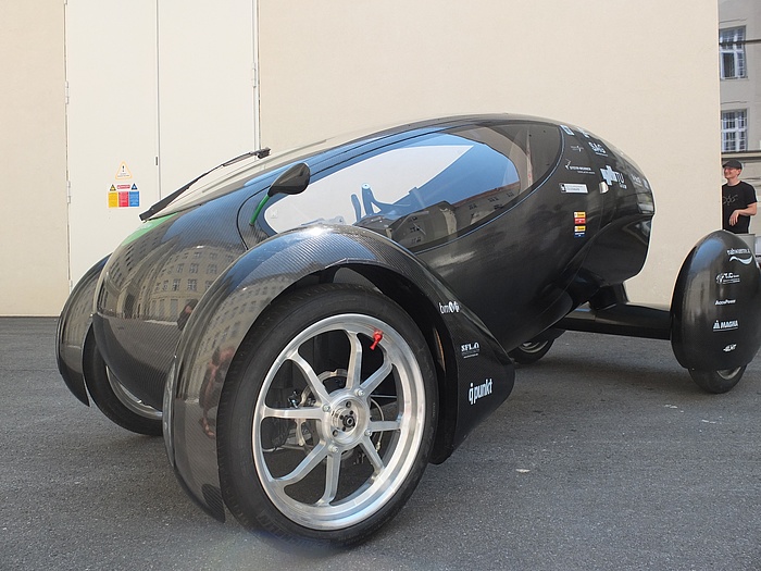 Small black egg-shaped vehicle, becoming increasingly narrower towards the end, with elegant, elongated oval windows, two big wheels at the front and a very wide wheel axle and smaller wheels at the rear.