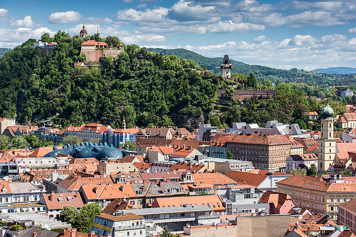 Blick auf die Dächerlandschaft des historischen Stadtkerns von Graz mit dem Schloßberg im Hintergrund.