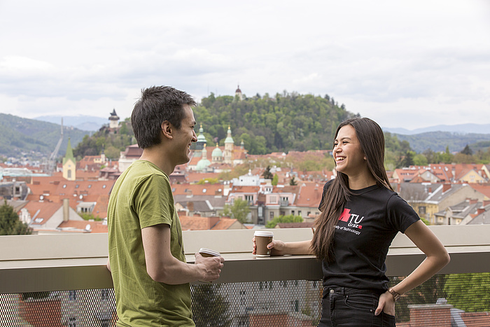 Ein Student im grünen T-Shirt und eine Studentin im schwarzen TU Graz-Shirt unterhalten sich bei einem Kaffee stehend auf einer Terrasse mit dem sommerlichen Grazer Schlossberg im Hintergrund.
