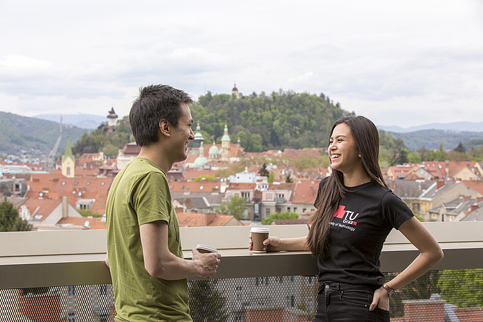 A male student in a green T-shirt and a female student in a black TU Graz shirt talk over coffee, standing on a terrace with a summery Graz Schlossberg in the background.