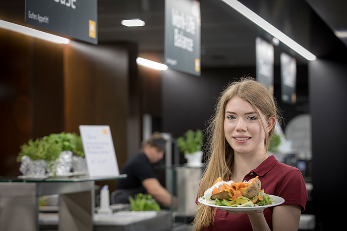 A female student wearing a wine-red polo shirt presents her plate of baked potatoes and vegetables in front of the service area in the Inffeldgasse canteen.