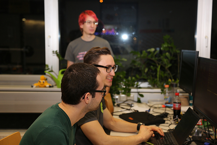 Zwei LosFuzzys-Jungs arbeiten im nächtlichen FuzzyLab gemeinsam am Computer, ein dritter steht daneben und schaut zu.
