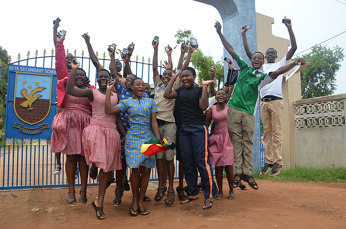 A group of students jumping up into the air in the open against the background of the iron access gate which bears the KETASCO logo.