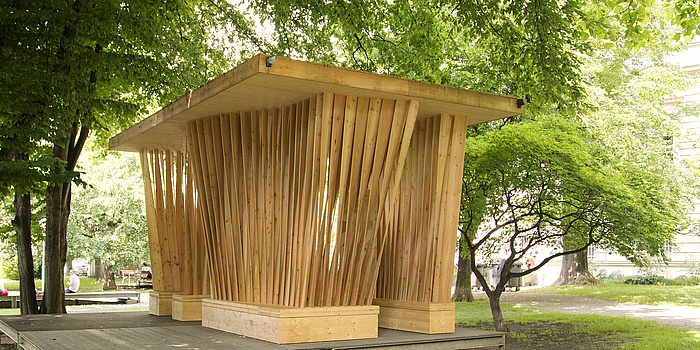 Wooden pavilion with vertical wooden-slat structure on a timber podium under a full-leafed tree in the park behind the Alte Technik.