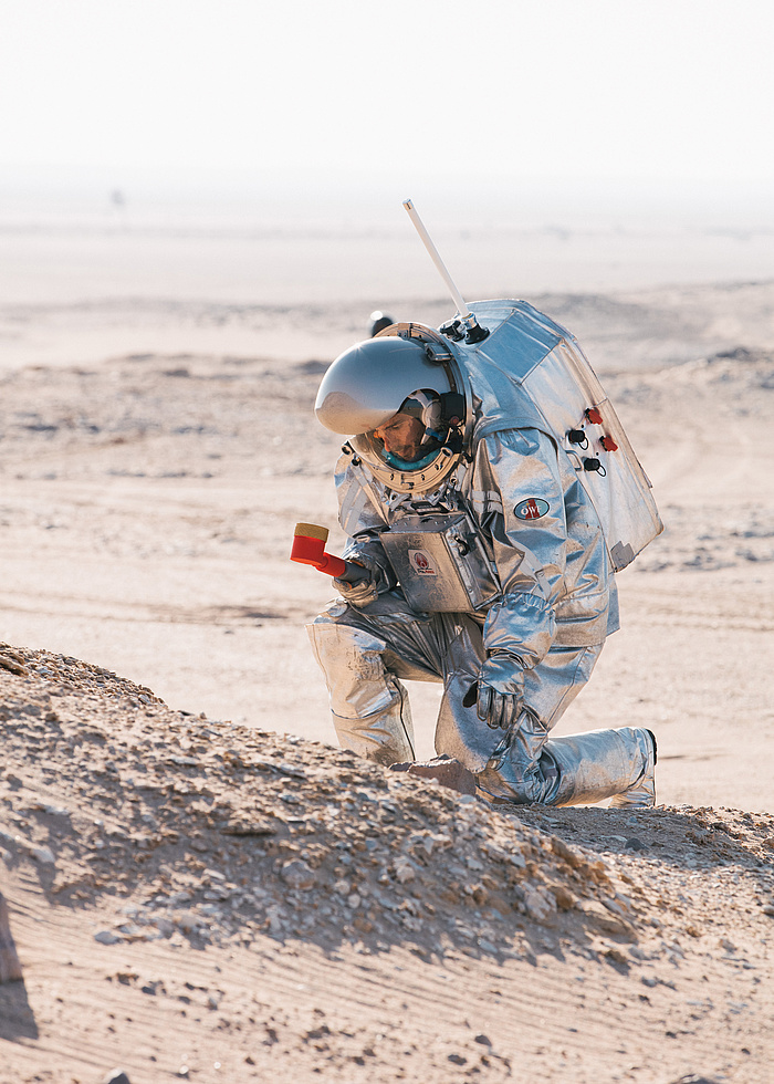 Ein Mann in einem RaumanzugA man in a space suit kneeling takes samples in the red desert sand. He holds a tool in one hand.