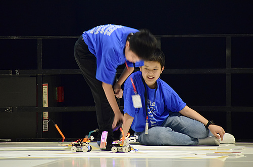 Zwei Jugendliche beobachten einen kleinen Roboter, der auf dem Boden entlangfährt.