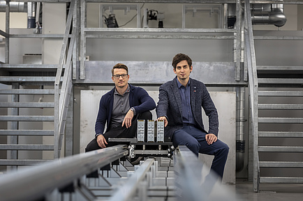 Two men sit on the rails of a test bench and look into the camera.