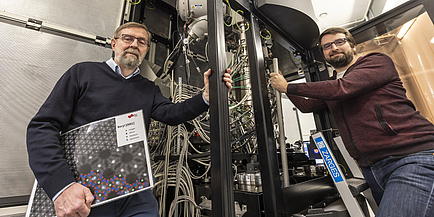 Two men stand next to an electron microscope and look into the camera.