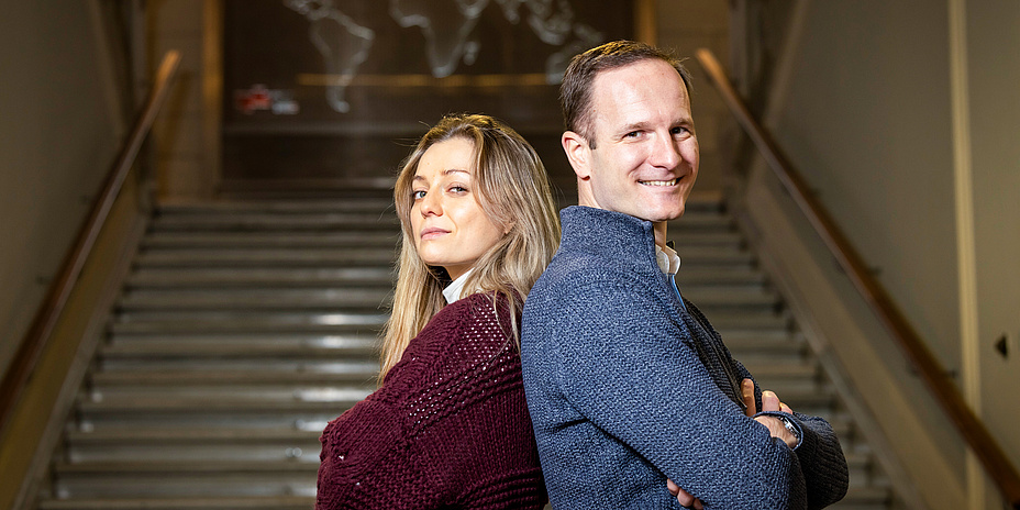 Ein Mann und eine Frau lehnen sich Rücken an Rücken und haben dabei die Arme verschränkt. Sie blicken selbstbewusst in die Kamera. 