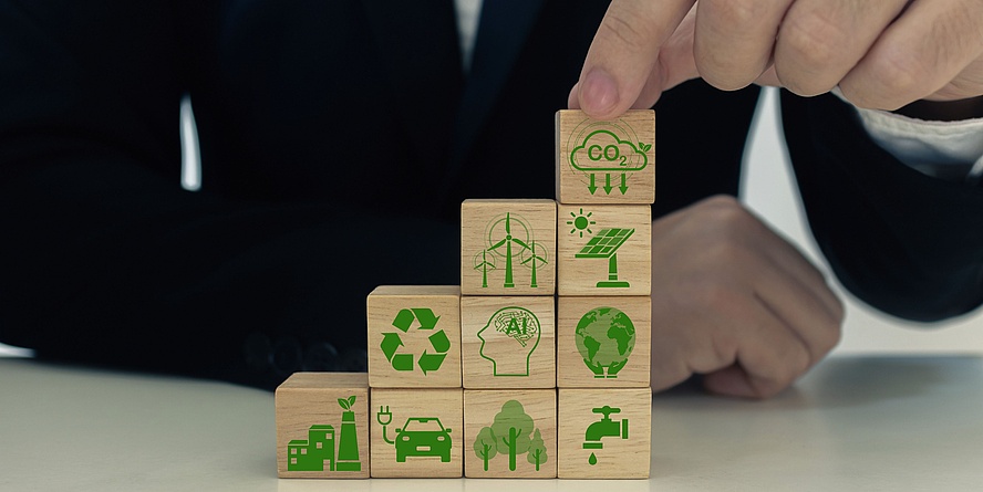 Person in black suit (cut-out torso and hands) stacks small wooden cubes with symbols for decarbonisation
