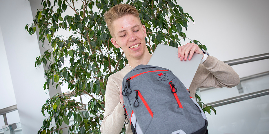 Young, fair-haired man in front of a big green-leafed plant puts documents into a grey-red rucksack.