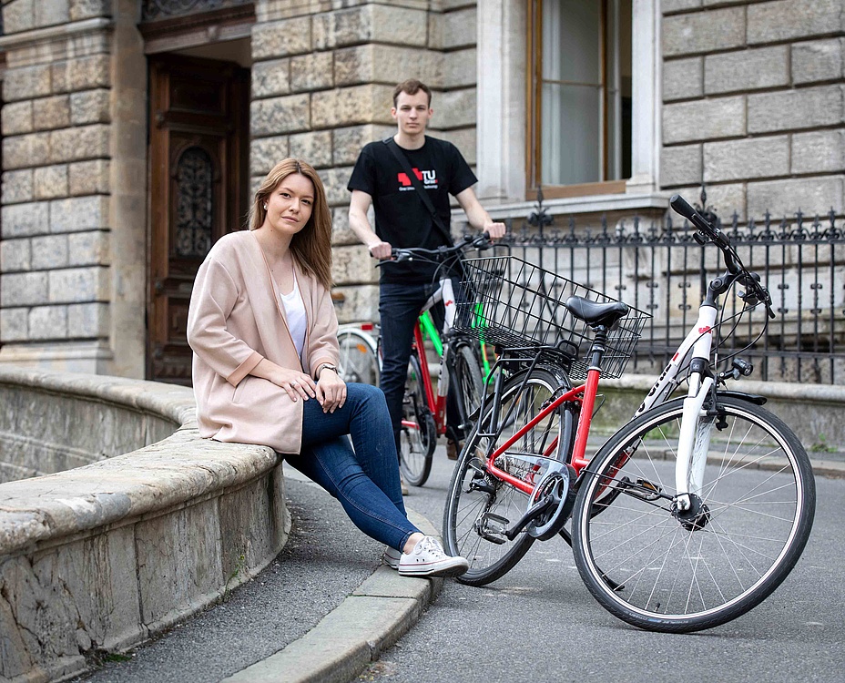 Junge Frau neben ihrem Fahrrad auf einer Steinmauer vor einem historischen Gebäude. Im Hintergrund sitzt ein junger Mann auf seinem Rad.