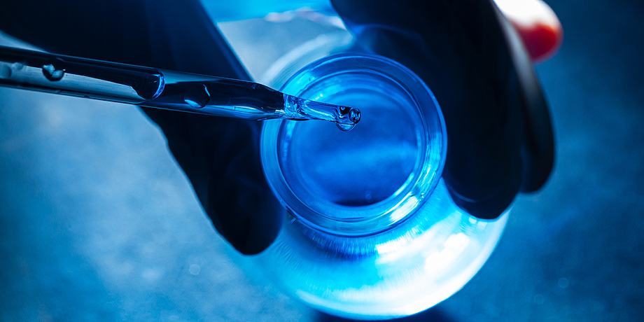 Droplets fall from a pipette into a glass container that is illuminated by blue light