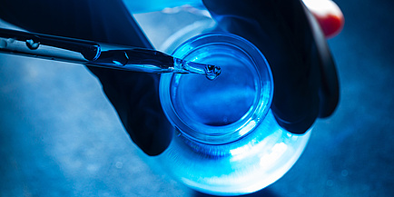 Droplets fall from a pipette into a glass container that is illuminated by blue light