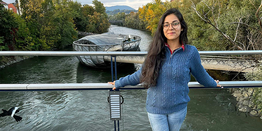 [Translate to Englisch:] Eine Frau mit langen, dunklen Haaren und Brille in blauer Weste und Jeans steht vor einem Brückengeländer. Hinter ihr fließt ein Fluss, darauf ist eine schwimmende Konstruktion zu sehen, die Murinsel. Links und rechts am Ufer stehen Bäume.