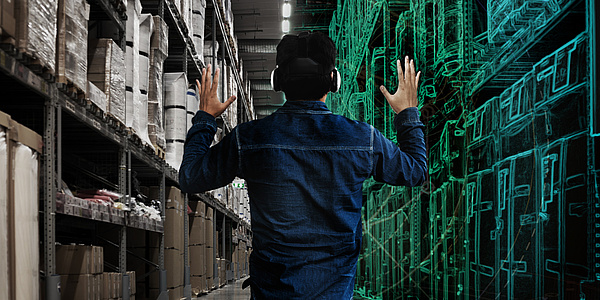 Rückansicht eines Mannes mit einer VR-Brille auf dem Kopf. Links von ihm ist ein Regal mit zahlreichen Kartons, rechts von ihm ein computergeneriertes Bild eines Regals.
