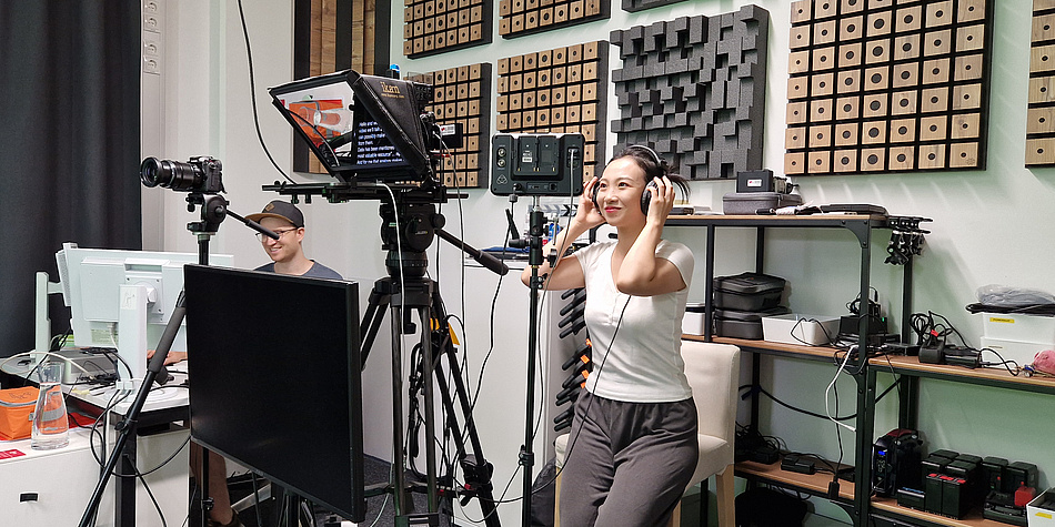 A Chinese woman in a white T-shirt sits on a stool, wears headphones and speaks into a microphone. 