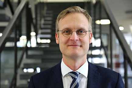 Ein blonder Mann mit Brille, Anzug und Krawatte steht vor einer Stiege und blickt in die Kamera