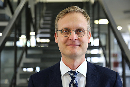 A blond man with glasses, suit and tie stands in front of a staircase and looks into the camera.