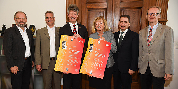 Stefan Vorbach, Dekan Roderick Bloem, Rektor Harald Kainz, Rektorin Christa Neuper, Dekan Thomas Foscht, Studiendekan Heinz Königsmaier stehen vor einer hölzernen Doppelflügeltüre. Rektor Harald Kainz und Rektorin Christa Neuper halten die A3-Werbeplakate in die Kamera. Bildquelle: Tzivanopoulos – UniGraz