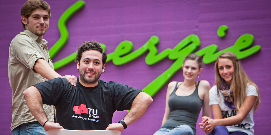 Two female and two male students of TU Graz in front of a bright green writing saying "Energie" in a building at Campus Inffeldgasse.