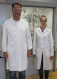 A man and a woman standing next to each other in a chemical laboratory wearing a white coat and goggles