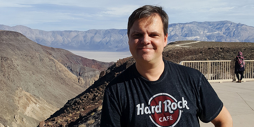 Ein Mann in einem schwarzen T-Shirt steht vor einer Landschaft aus sanften Hügeln.