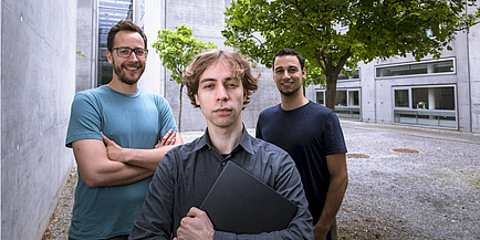 Daniel Gruss in the front carries a closed laptop. His hair is blond and he is wearing a black shirt. On his left is Moritz Lipp, who wears dark hair and a blue shirt. On his right is Michael Schwarz, who wears dark hair and a black shirt.