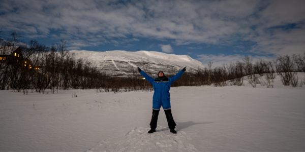 Ein Studierender im Schneeanzug steht mit weit ausgebreiteten Armen in der schneebedeckten Landschaft.