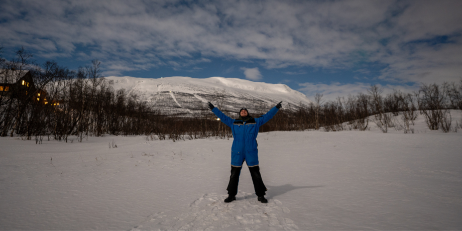 Ein Studierender im Schneeanzug steht mit weit ausgebreiteten Armen in der schneebedeckten Landschaft.