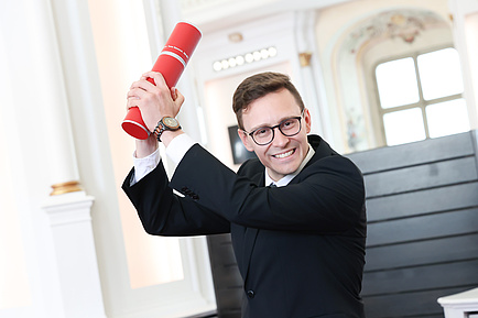 A man holds up a red document roll and smiles into the camera.