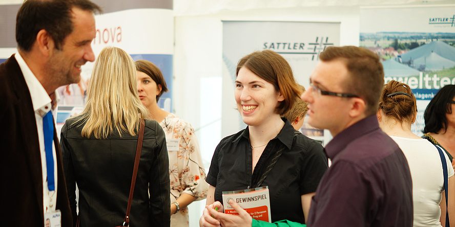 A young man and woman talking to a middle-aged representative with a tie at TECONOMY Graz. In the background, more visitors in front of company information stands.