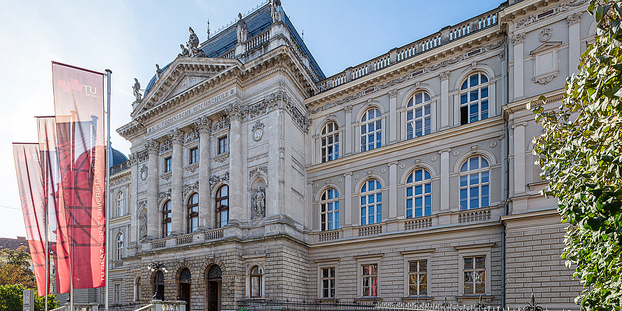 A large historical building, in front of which flags in TU Graz look are hoisted.