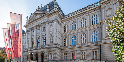 Ein großes historisches Gebäude, davor sind Fahnen in TU Graz-Optik gehisst.