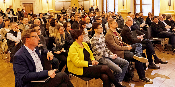 In einer großen Aula sind voll besetzte Stuhlreihen zu sehen.