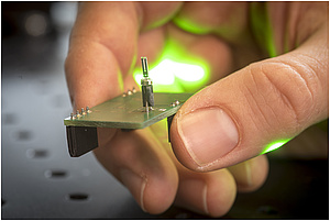 A hand holds a small circuit board with a tuning fork attachted to it