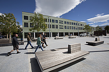 Multi-storey green building, people walk across a square in front of it.