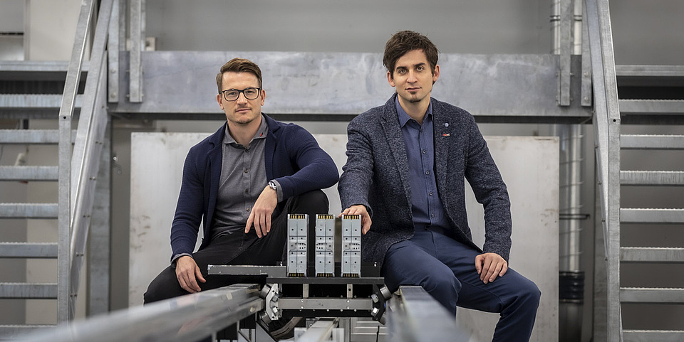 Two men sit next to each other on the rails of a test bench and look into the camera.