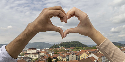 Blick auf das historische Zentrum einer Stadt mit Berglandschaft im Hintergrund und zwei Händen, die ein Herz formen, im Vordergrund.