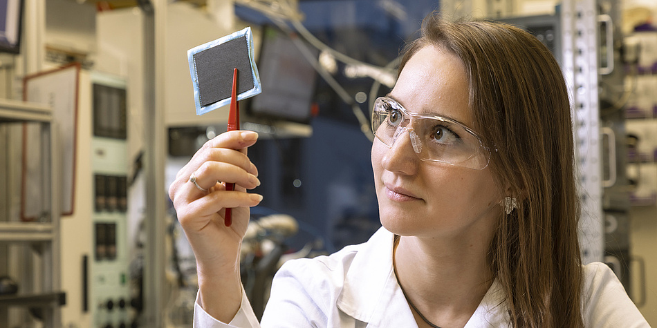 A young woman with long brown hair is wearing a white lab coat and protective goggles. In her hand, she holds a black, square object with red tweezers.